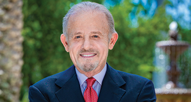 Headshot of a man with dark suit and red tie outside in front of fountain