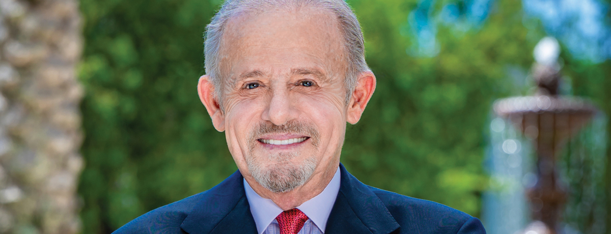 Headshot of a man with dark suit and red tie outside in front of fountain