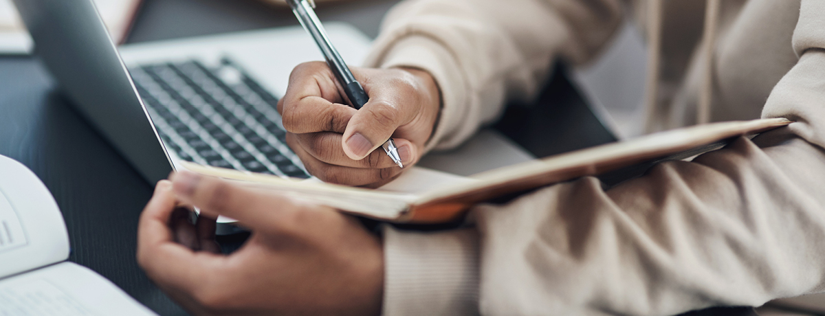 Person taking notes in journal with open laptop in background