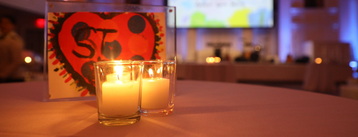 Two candles on table in front of framed child's artwork and large screen at event