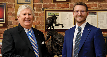 Two lawyers in suits standing in front of brick wall with awards