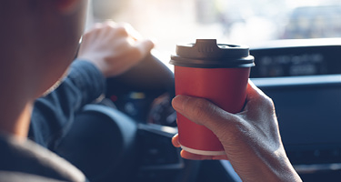 Man with hand on the steering wheel drinks and holds red cup of coffee with a black lid