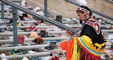 Indigenous woman stands near piles of children's shoes 