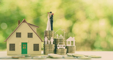 Toy house and figure of married couple standing on stacks of coins