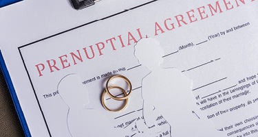 Two gold rings and paper cutouts of a couple over prenup papers on clipboard