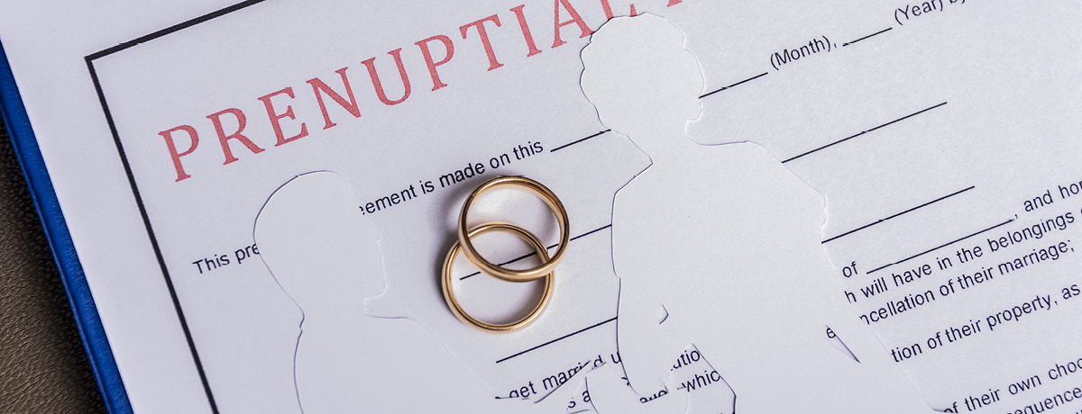 Two gold rings and paper cutouts of a couple over prenup papers on clipboard