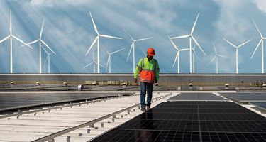 Man with construction helmet inspecting solar panels with wind turbines in backdrop