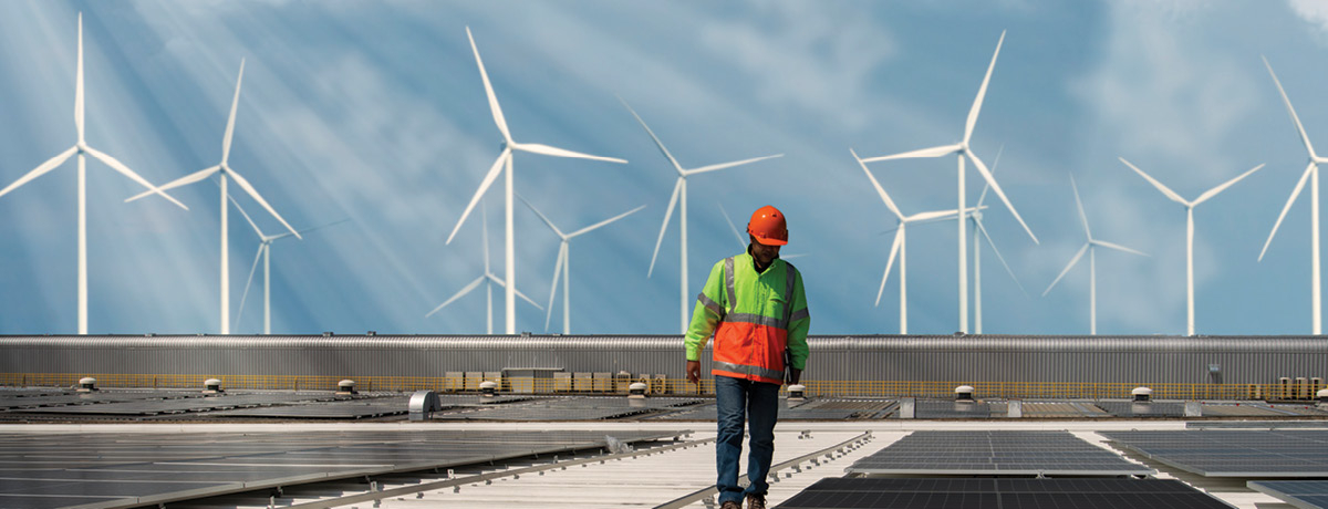 Man with construction helmet inspecting solar panels with wind turbines in backdrop