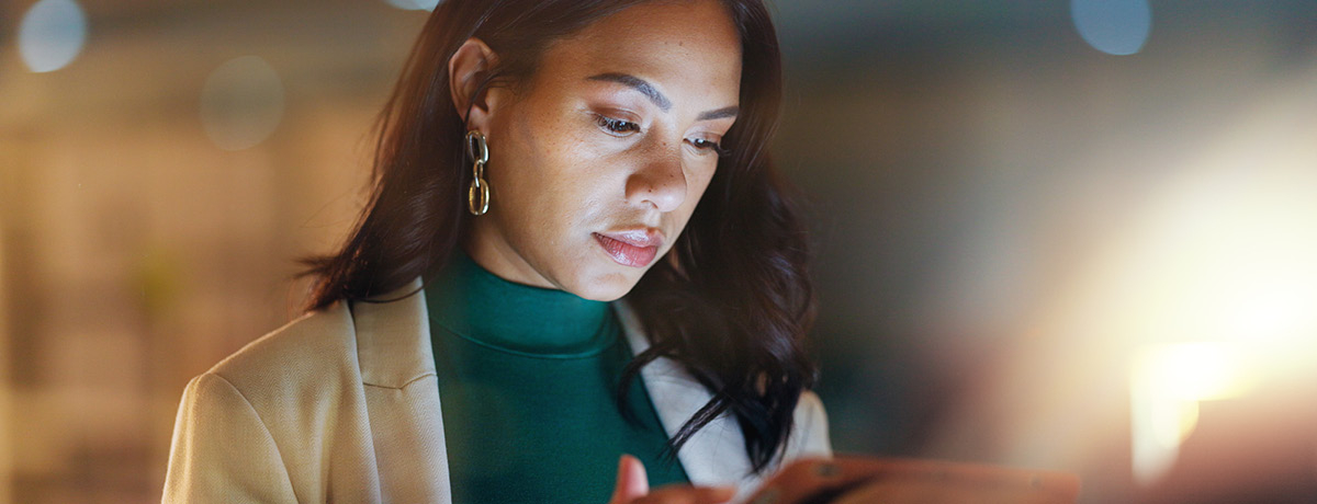A focused woman with dark hair wearing a green top and beige blazer, working on a tablet in a dimly