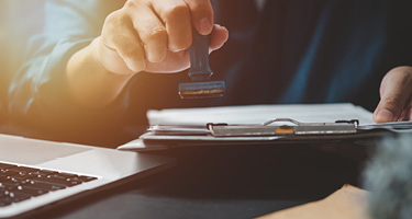 A White Hand Holding a Stamp Above Papers on a Clipboard in Front of a Laptop