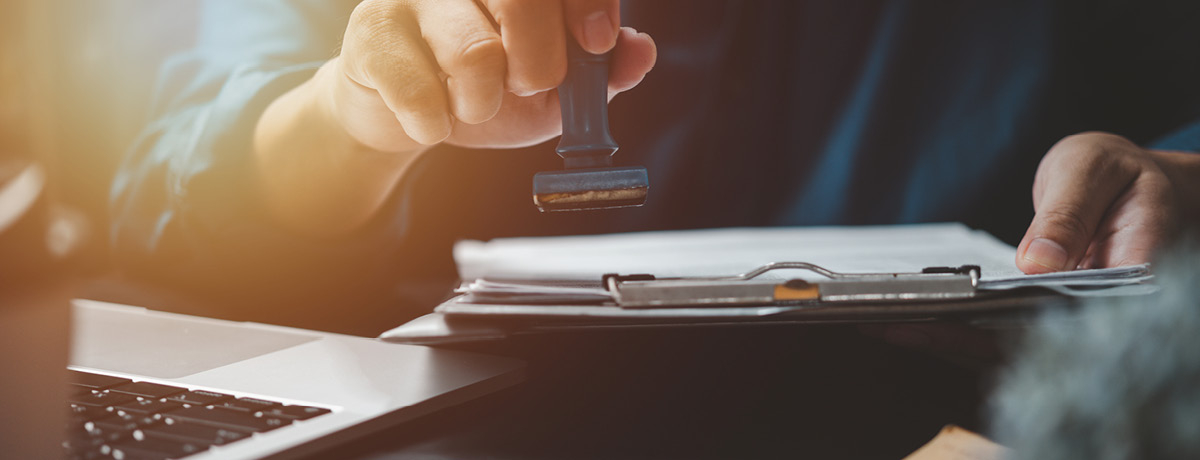A White Hand Holding a Stamp Above Papers on a Clipboard in Front of a Laptop