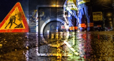 Road at night with two people wearing blue suits and a red triangle warning sign with a magnifying g