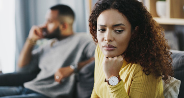 Woman sitting on couch rests head on fist with man in background