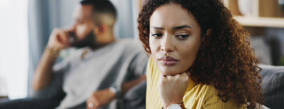 Woman sitting on couch rests head on fist with man in background