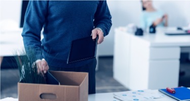Office with a man wearing a blue sweater holding a tablet and packing his things