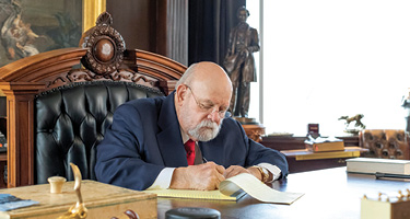 Suited man sits behind desk and writes on notepad