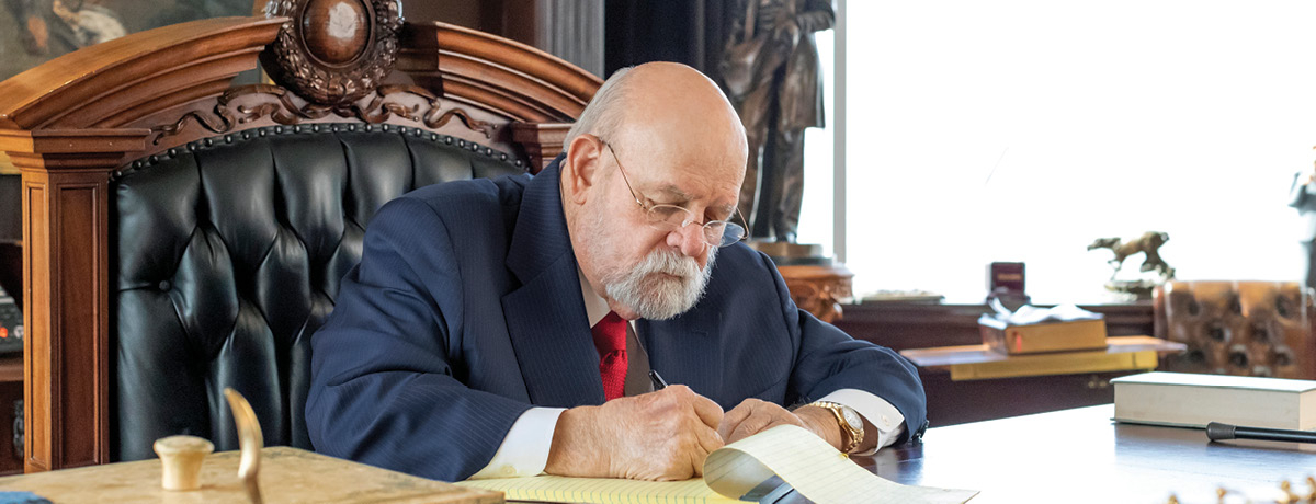 Suited man sits behind desk and writes on notepad