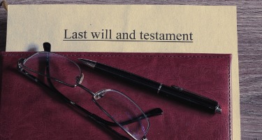 Pair of reading glasses, a pen, and a folder or file on a wooden desk.