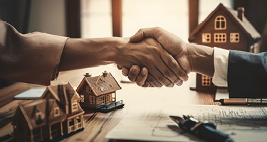 Two unseen individuals shake hands with miniature house models in background
