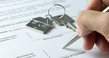 Individual signing a document with house keys in background