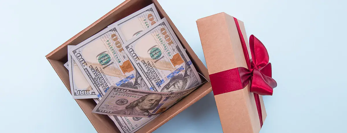 An open gift box filled with US $100 bills next to a lid tied with a red bow.