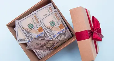 An open gift box filled with US $100 bills next to a lid tied with a red bow.