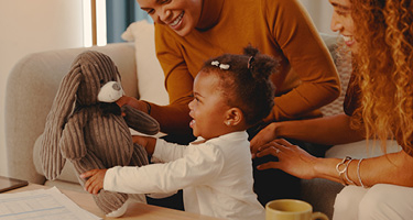 Child holding a teddy bear with two women sitting with her