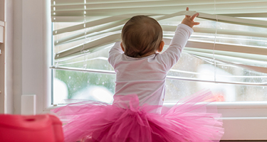 Baby wearing pink tutu playing with window blinds
