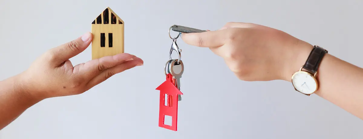 One hand holding a wooden house model and another holding house keys.