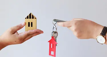 One hand holding a wooden house model and another holding house keys.